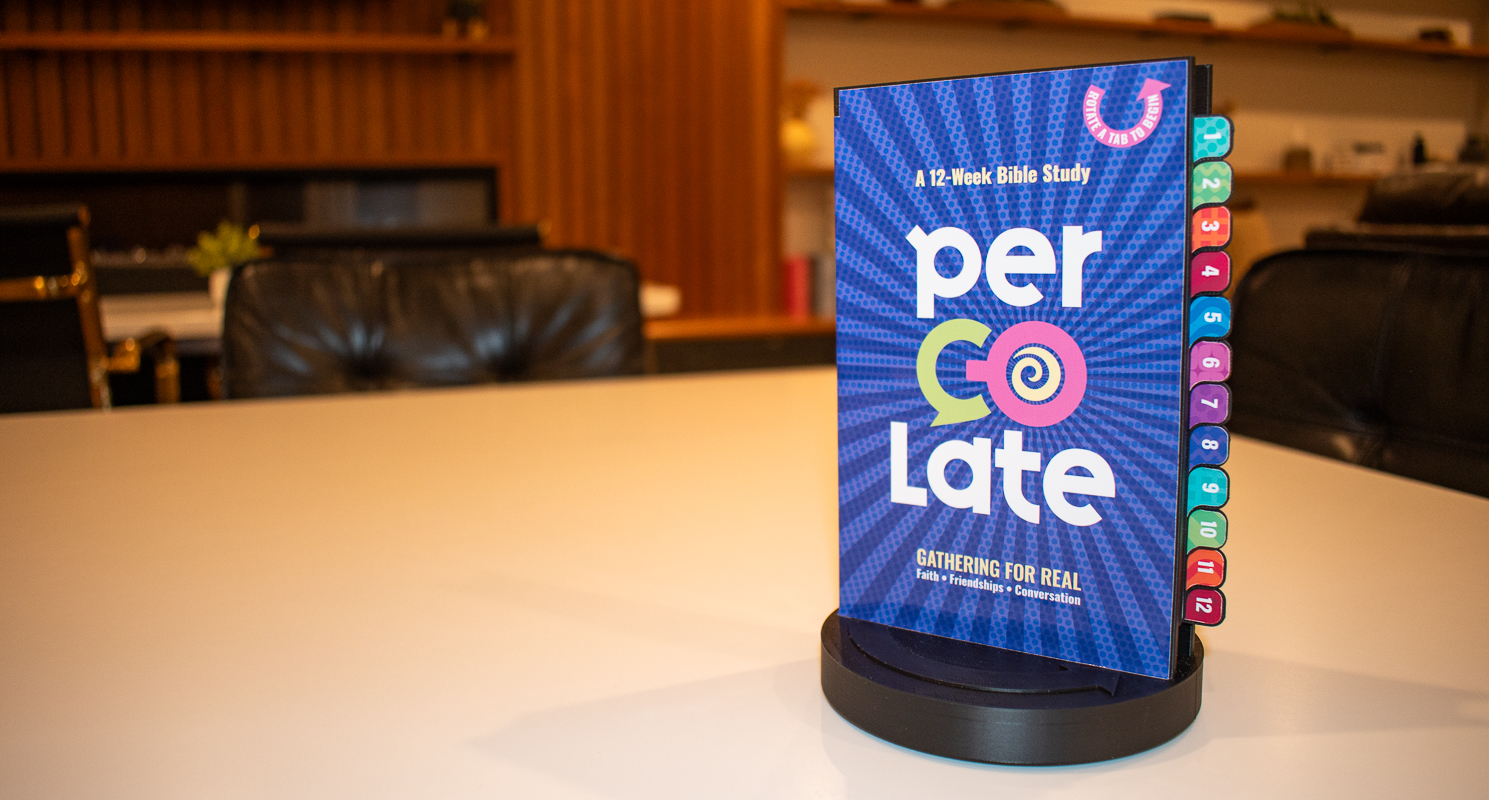 Percolate Bible Study stand on a coffee shop table — a book-shaped 3D-printed stand on a round spinning base with 12 colorful numbered tabs visible on the side